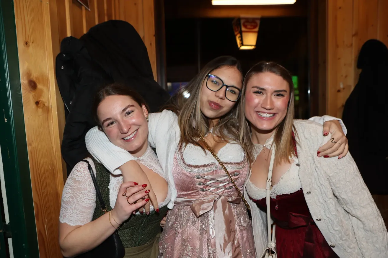 Drei Frauen lächeln und posieren für ein Foto. Die Frau auf der linken Seite trägt einen grünen Pullover und eine schwarze Tasche. Die Frau in der Mitte trägt einen weißen Pullover und eine Brille. Die Frau auf der rechten Seite trägt ein rotes Kleid.