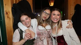 Drei Frauen lächeln und posieren für ein Foto. Die Frau auf der linken Seite trägt einen grünen Pullover und eine schwarze Tasche. Die Frau in der Mitte trägt einen weißen Pullover und eine Brille. Die Frau auf der rechten Seite trägt ein rotes Kleid.