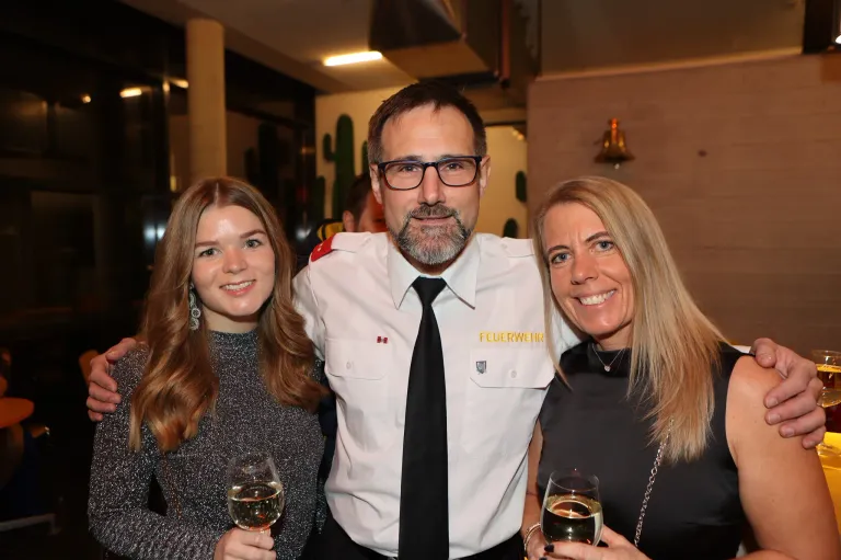 Drei Menschen lächeln und posieren für ein Foto. Der Mann trägt eine weiße Uniform mit einer Namensplakette, die 'Feuerwehr' besagt. Die Frau auf der linken Seite hält ein Glas Wein. Die Frau auf der rechten Seite hält ebenfalls ein Glas Wein. Sie stehen eng beieinander. Hinter ihnen ist ein verschwommenes Bild einer Wand mit einer Glocke und einer Kaktusdekoration.