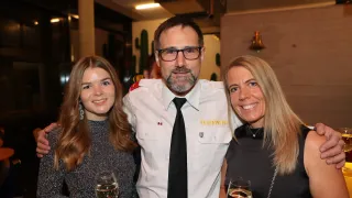 Drei Menschen lächeln und posieren für ein Foto. Der Mann trägt eine weiße Uniform mit einer Namensplakette, die 'Feuerwehr' besagt. Die Frau auf der linken Seite hält ein Glas Wein. Die Frau auf der rechten Seite hält ebenfalls ein Glas Wein. Sie stehen eng beieinander. Hinter ihnen ist ein verschwommenes Bild einer Wand mit einer Glocke und einer Kaktusdekoration.