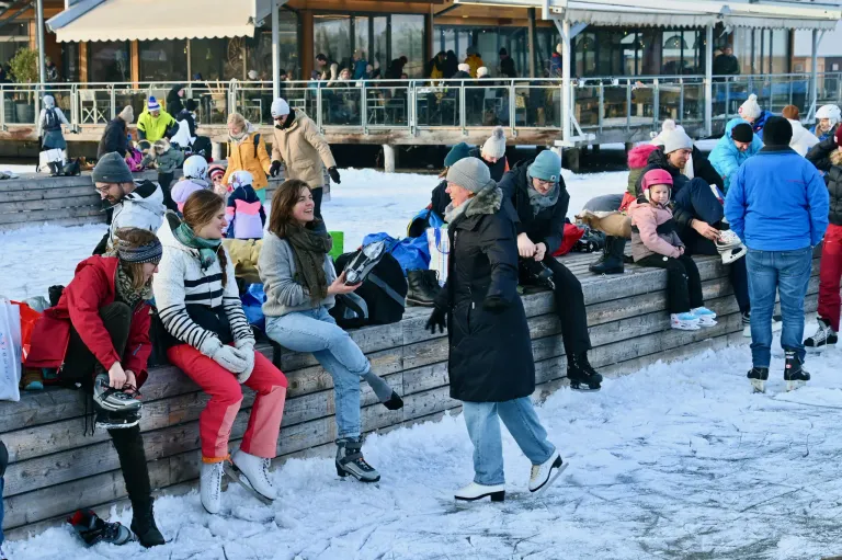 Eine Gruppe von Menschen sitzt an einem verschneiten Tag auf einer Holzmauer. Einige tragen Eislaufschuhe. Sie lächeln und plaudern. Hinter ihnen ist ein Gebäude mit Glaswänden zu sehen.