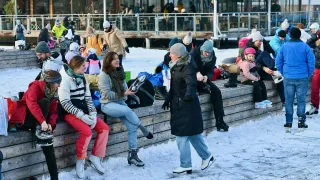 Eine Gruppe von Menschen sitzt an einem verschneiten Tag auf einer Holzmauer. Einige tragen Eislaufschuhe. Sie lächeln und plaudern. Hinter ihnen ist ein Gebäude mit Glaswänden zu sehen.