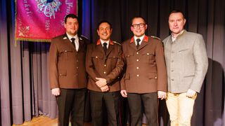 Vier Männer in braunen Uniformen stehen vor einem schwarzen Vorhang, lächeln und posieren für ein Foto. Der Mann auf der linken Seite trägt eine Armbanduhr.
