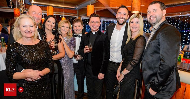 Mehrere Personen in formeller Kleidung posieren für ein Foto, Getränke in der Hand, in einem Raum mit warmer Beleuchtung und Weihnachtsdekoration.