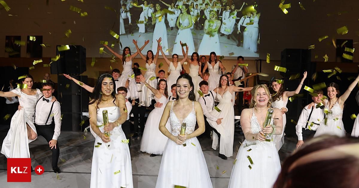 Eine Gruppe von Menschen in weißen Kleidern wirft Konfetti auf einer Bühne, mit einer Projektionsleinwand dahinter, die eine Menschenmenge zeigt.