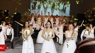 Eine Gruppe von Menschen in weißen Kleidern wirft Konfetti auf einer Bühne, mit einer Projektionsleinwand dahinter, die eine Menschenmenge zeigt.