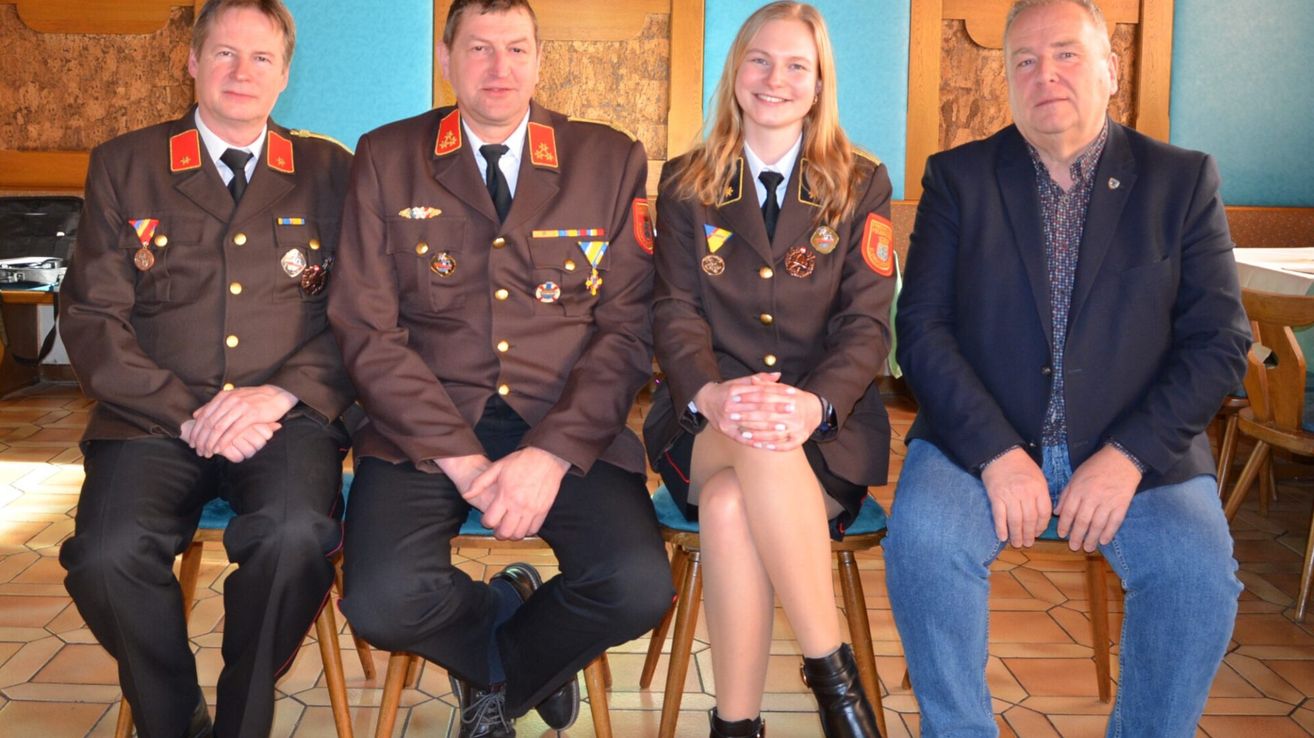 Drei Menschen in Uniform, zwei Männer und eine Frau, sitzen auf Stühlen und lächeln für ein Foto. Der Mann auf der linken Seite trägt eine braune Uniform mit Medaillen. Die Frau hat blondes Haar und trägt eine braune Uniform mit Abzeichen. Der Mann auf der rechten Seite trägt einen blauen Anzug.