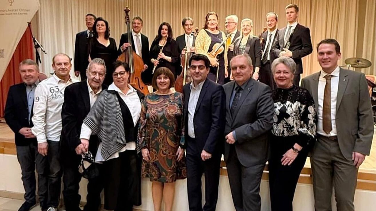 Eine Gruppe von Musikern, darunter Streicher, Bläser und Blechbläser, stehen zusammen, einige halten ihre Instrumente. Sie scheinen bei einer formellen Veranstaltung zu sein, mit Männern in Anzügen und Frauen in eleganten Kleidern.