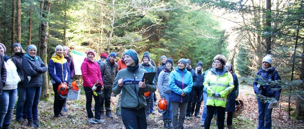 Eine Gruppe von Menschen in Winterkleidung und Helmen steht in einem Wald, wobei eine Person ein Klemmbrett hält und spricht.