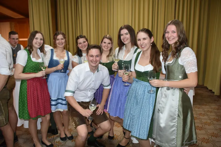 Eine Gruppe lächelnder Frauen und ein Mann in traditioneller bayerischer Kleidung posiert zusammen und hält Weingläser. Sie scheinen bei einer Veranstaltung zu feiern.