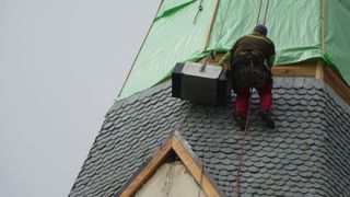 Ein Arbeiter in Sicherheitsausrüstung klettert auf ein Dach und hält eine große Kiste. Grüne Planen bedecken den oberen Teil des Daches.