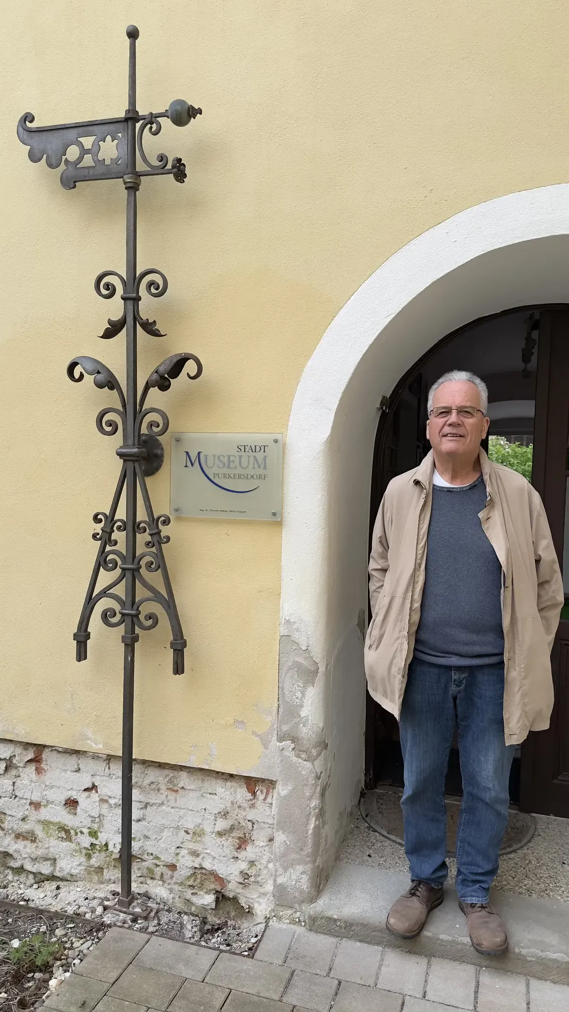 Ein älterer Mann mit Brille steht vor einer Tür, die mit Stadt Museum Purkersdorf beschriftet ist. Er trägt eine leichte Jacke und blaue Jeans. Ein dekorativer Metallpfosten ist an der Wand angebracht.