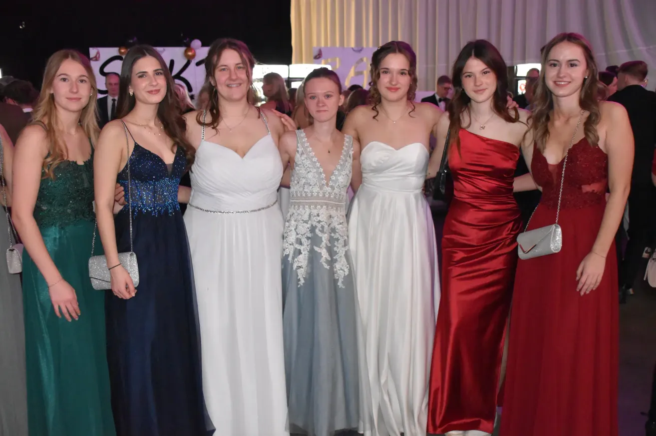 Fünf Frauen stehen bei einer formellen Veranstaltung zusammen. Sie tragen elegante Kleider und lächeln für ein Foto. Die Frau links trägt ein blaues Kleid.