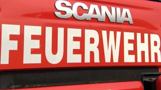 Ein rotes Schild mit weißen Buchstaben, die 'SCANIA' und 'FEUERWEHR' lesen, ist auf einem roten Hintergrund angebracht. Die Buchstaben sind groß und fett, und das Schild ist wahrscheinlich an einem Fahrzeug befestigt.