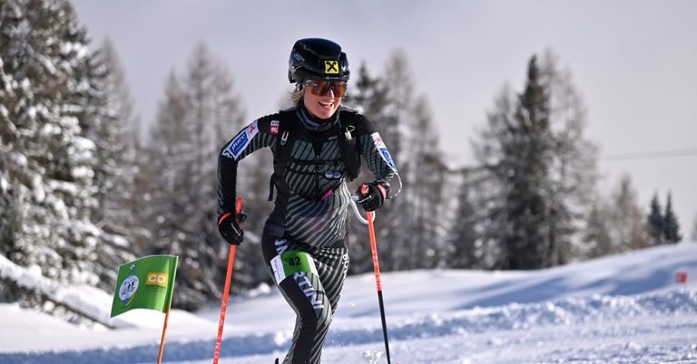 Eine Skifahrerin nimmt an einem Langlaufrennen teil, trägt einen Helm, eine Schutzbrille und Skiausrüstung. Sie fährt auf einem verschneiten Feld mit Tannen im Hintergrund.