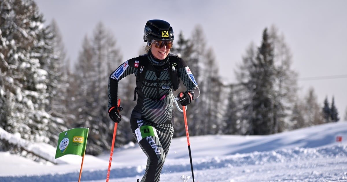Eine Skifahrerin nimmt an einem Langlaufrennen teil, trägt einen Helm, eine Schutzbrille und Skiausrüstung. Sie fährt auf einem verschneiten Feld mit Tannen im Hintergrund.
