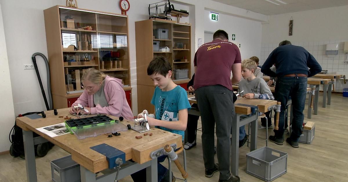 Eine Gruppe von Kindern und Erwachsenen arbeitet in einer Werkstatt an einem Projekt. Sie sind von Holztischen und Stühlen umgeben, und Werkzeuge und Materialien sind auf den Tischen verstreut.