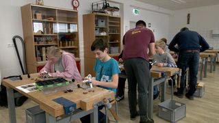 Eine Gruppe von Kindern und Erwachsenen arbeitet in einer Werkstatt an einem Projekt. Sie sind von Holztischen und Stühlen umgeben, und Werkzeuge und Materialien sind auf den Tischen verstreut.