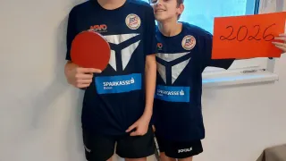 Zwei Jungen stehen lächelnd in der Nähe eines Fensters und halten Schläger. Ein Junge hält einen roten Schläger und ein Schild mit 2026. Sie tragen passende blaue Trikots mit weißen Akzenten und schwarze Shorts. Ein Fenster mit Blick auf ein Gebäude befindet sich hinter ihnen.