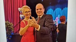 Ein Mann und eine Frau lächeln und tanzen bei einer formellen Veranstaltung. Die Frau trägt ein rotes Kleid und der Mann einen Anzug. Sie sind von anderen Leuten umgeben, und im Hintergrund befindet sich eine Bühne mit Luftballons und blauen Lichtern.