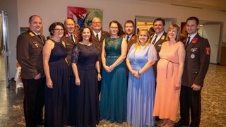 Eine Gruppe von Personen, sowohl Männer als auch Frauen, sind feierlich gekleidet und lächeln für ein Foto. Einige tragen Uniformen, und im Hintergrund sind ein Gemälde und eine Tür zu sehen.