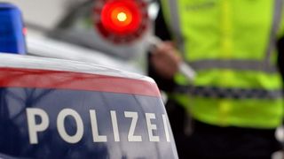 Ein Polizeibeamter in einem gelben Weste steht neben einem Polizeiwagen, das Wort 'POLIZEI' ist auf dem Auto sichtbar.