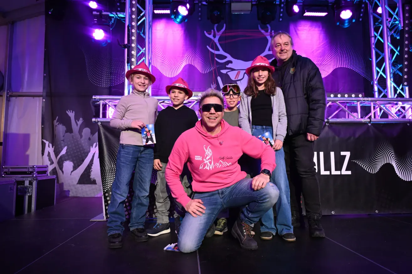 Eine Gruppe von Kindern und Erwachsenen auf einer Bühne, alle lächeln und posieren für ein Foto. Sie sind in Winterkleidung gekleidet und tragen Hüte. Hinter ihnen ist ein Hintergrund mit einem Hirschdesign.