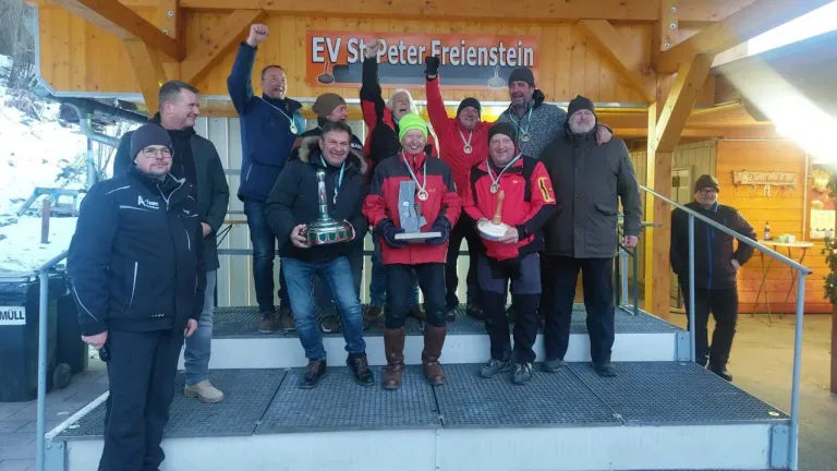 Gruppe von Männern feiert auf einer Bühne, hält Trophäen in den Händen, mit einem Banner hinter ihnen, das 'EV St Peter Freienstein' steht.