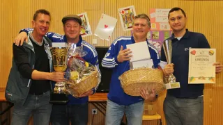 Vier Männer posieren mit Trophäen und Geschenken für ein Foto. Sie lächeln und scheinen einen Sieg zu feiern.