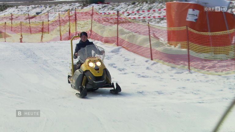 Eine Person fährt auf einer verschneiten Strecke mit einem Schneemobil, hinter ihr befindet sich ein roter Sicherheitszaun. Sie trägt eine schwarze Jacke und einen schwarzen Hut.