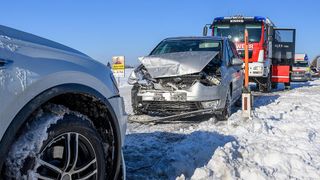 Ein silberner Wagen mit beschädigtem vorderen Stoßfänger steht im Schnee. Ein roter Feuerwehrwagen ist in der Nähe. Ein Schild mit einem Bild eines Mannes steht am Straßenrand.