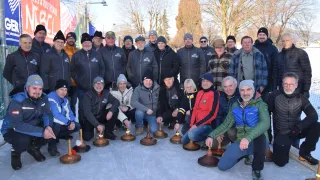 Eine Gruppe von Erwachsenen in Winterkleidung posiert für ein Foto auf Eis. Sie sitzen und knien mit Objekten vor sich. Hinter ihnen stehen Bäume und eine Straßenlaterne.