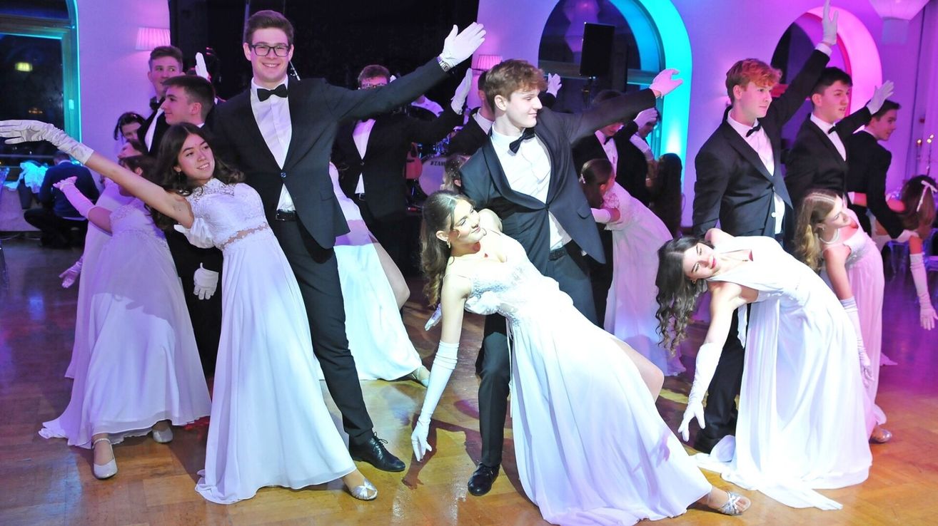 Eine Gruppe von Menschen in formeller Kleidung tanzt auf einem Holzboden unter lila Beleuchtung. Eine Frau in einem weißen Kleid wird von einem Mann in einem Smoking herumgedreht.