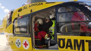 Ein Rettungshubschrauber mit offener Tür, zwei Personen im Inneren in Schutzausrüstung, rotem Kreuz an der Seite und den Buchstaben OA an der Seite der Tür.