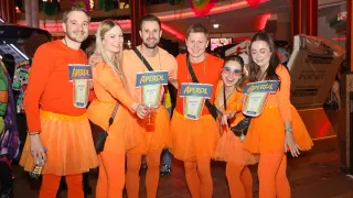 Eine Gruppe von Menschen in orangefarbenen Kostümen posiert für ein Foto mit Aperol-Schildern bei einer Veranstaltung. Der Hintergrund zeigt eine lebhafte Indoor-Party mit Dekorationen und anderen Menschen.