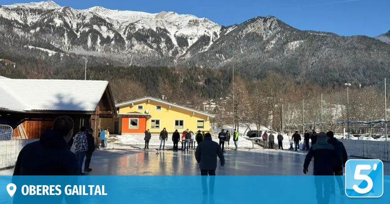 Eine Gruppe von Menschen Schlittschuh fährt auf einer Eisbahn in einem verschneiten Gebirgsgebiet mit einem gelben Gebäude hinter ihnen.