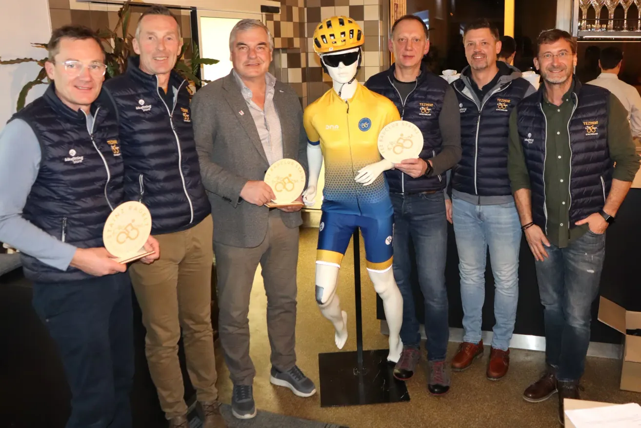 Fünf Männer stehen neben einem Mannequin in Radfahrer-Ausrüstung und halten Medaillen. Das Mannequin trägt ein gelbes und blaues Radtrikot. Die Männer tragen Jacken mit Logos und Nummern.