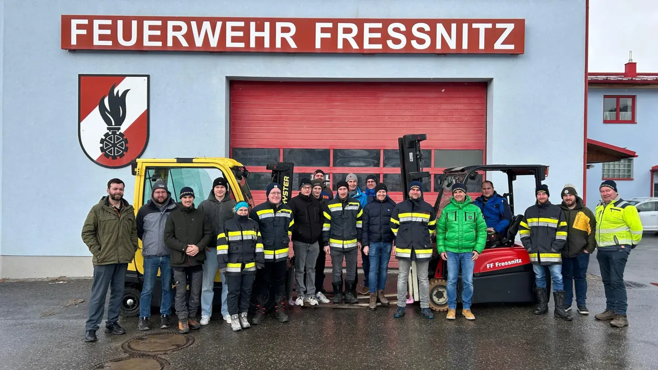 Eine Gruppe von Menschen in Winterkleidung steht vor einer roten Garage mit dem Schild Wehr Wehr Fressnitz. Einige Leute tragen Mützen und andere Stiefel. Zwei Gabelstapler sind vor der Garage geparkt.