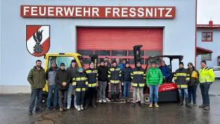 Eine Gruppe von Menschen in Winterkleidung steht vor einer roten Garage mit dem Schild Wehr Wehr Fressnitz. Einige Leute tragen Mützen und andere Stiefel. Zwei Gabelstapler sind vor der Garage geparkt.