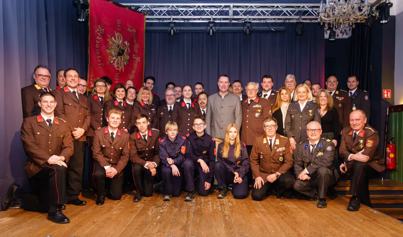 Eine Gruppe von Menschen in Militäruniformen posiert für ein Foto auf einer Bühne mit einem blauen Vorhang und einem roten Banner im Hintergrund.