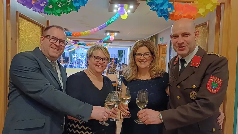 Drei Menschen, die Weingläser halten, zwei Frauen und ein Mann, lächeln in einem Raum, in dem eine Band spielt. Der Raum ist mit Dekorationen an der Decke geschmückt.