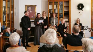 Mehrere Personen versammeln sich in einem Raum mit einem Kamin. Eine Frau steht vor einem Mikrofon und hält ein Papier in der Hand. Hinter ihr steht ein Mann neben einem Porträt an der Wand. Ein Violinist sitzt auf einem Stuhl. Ein Bücherregal voller Bücher befindet sich rechts.