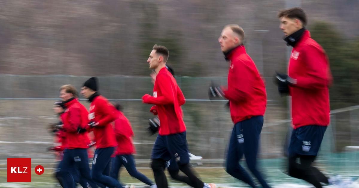 Eine Gruppe von Männern in roten Trikots und blauen Shorts läuft auf einem Feld. Der Mann vorne ist verschwommen, und die anderen sind leicht verschwommen. Sie tragen alle Handschuhe und Turnschuhe. Dahinter ist ein verschwommener Blick auf einen Zaun und Bäume.