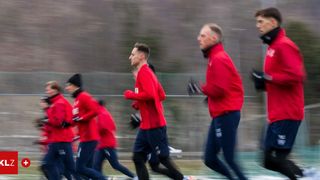 Eine Gruppe von Männern in roten Trikots und blauen Shorts läuft auf einem Feld. Der Mann vorne ist verschwommen, und die anderen sind leicht verschwommen. Sie tragen alle Handschuhe und Turnschuhe. Dahinter ist ein verschwommener Blick auf einen Zaun und Bäume.