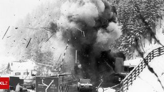 Ein Schwarz-Weiß-Foto zeigt eine Fahrzeugexplosion mit fliegenden Trümmern. Ein Auto ist zerschossen, und Rauch steigt vor einem Straßenschild auf. Bäume und ein Gebäude sind im Hintergrund.