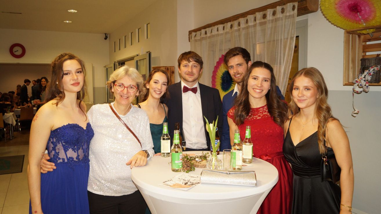 Eine Gruppe von Menschen versammelt sich um einen runden Tisch, lächelt und posiert für ein Foto. Zwei Männer sind in Anzügen gekleidet, der linke trägt eine Fliege. Drei Frauen stehen um den Tisch, eine mit einem roten Kleid und eine andere mit einem silbernen Kleid.