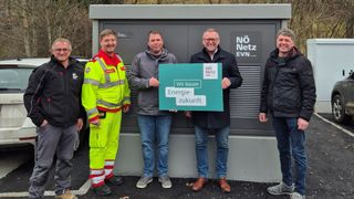 Drei Männer stehen vor einem Kraftwerk. Der Mann auf der linken Seite trägt eine reflektierende Warnweste, der Mann in der Mitte eine graue Jacke und der Mann auf der rechten Seite einen schwarzen Mantel. Sie halten ein grünes Schild mit der Aufschrift 'Wir bauen Energie-zukunft.' Hinter ihnen steht ein Gebäude mit einem Schild, auf dem 'Nö Netz EVN' steht.