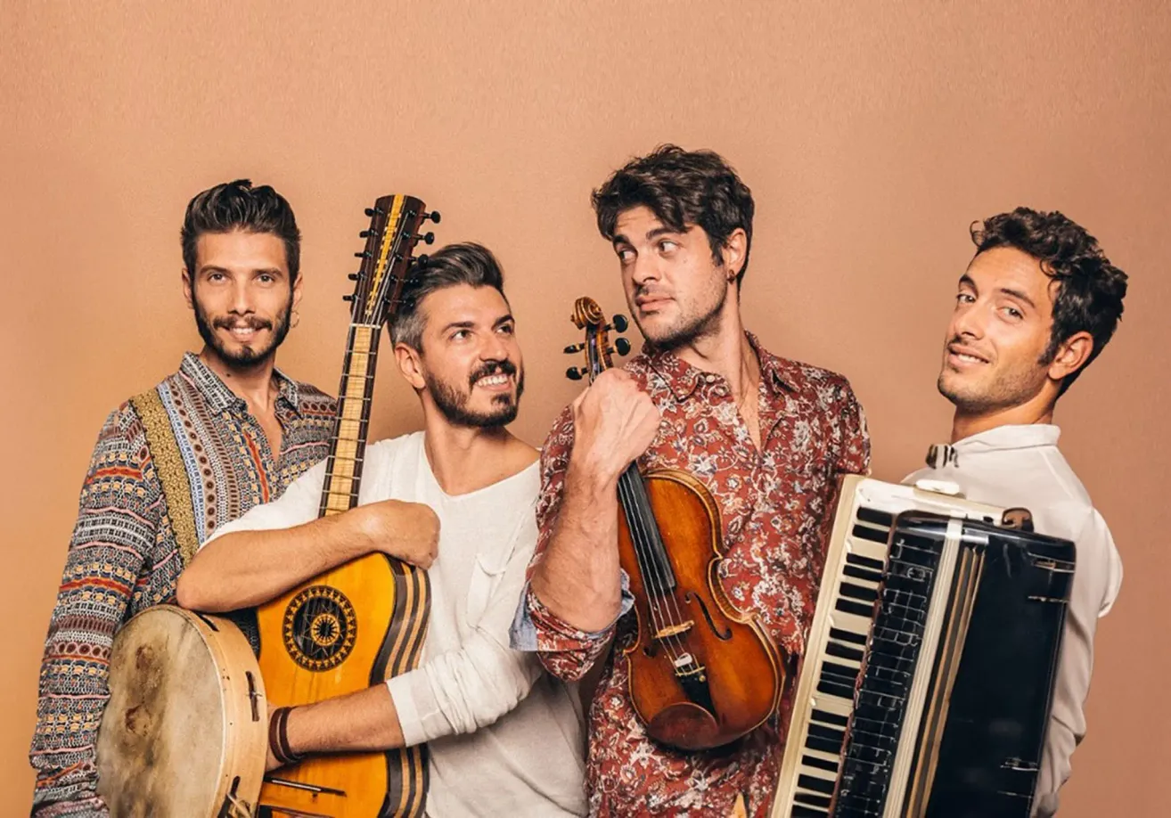 Fünf Männer stehen und posieren für ein Foto. Ein Mann auf der linken Seite hält eine Gitarre und eine Trommel. Ein anderer Mann in der Mitte hält eine Geige. Ein Mann auf der rechten Seite hält ein Akkordeon. Die beiden Männer an der Seite lächeln.
