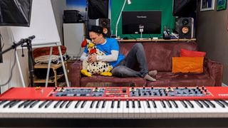Ein Mann in einem blauen Shirt und Hut sitzt auf einem Sofa mit einem Stoffhund. Er ist von Musikequipment umgeben, einschließlich eines Keyboards und Lautsprecher.