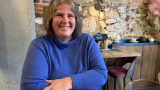 Eine Frau in einem blauen Pullover lächelt in einem Restaurant mit einer Steinmauer in die Kamera.